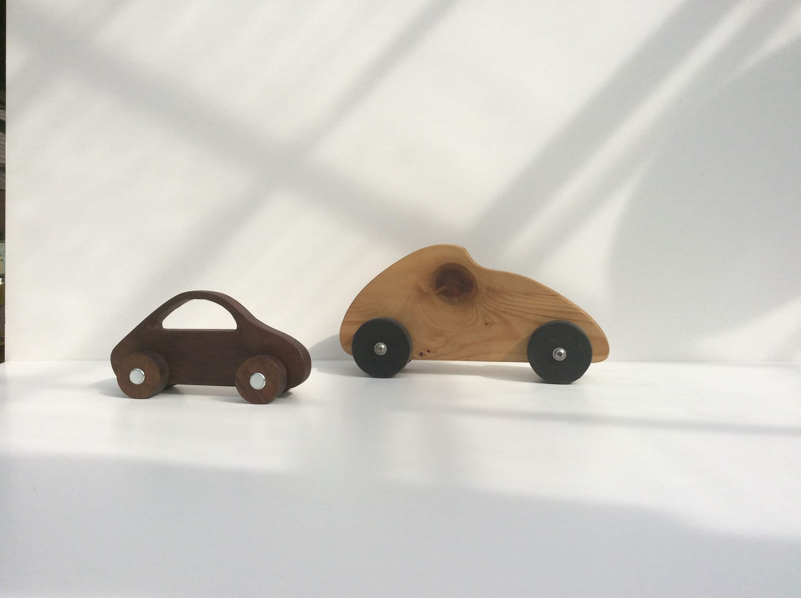 Twee houten speelgoedautootjes naast elkaar op een witte tafel, in het strijklicht.