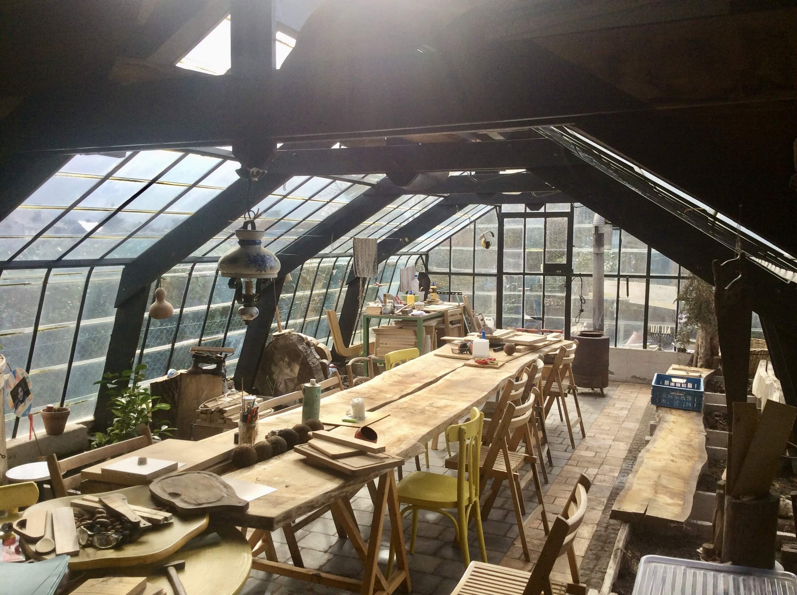 De serre van Bert Lemmens: een lange werkbank van ruwe planken onder een glazen kap, met houten stoelen, gereedschap en werk in uitvoering.