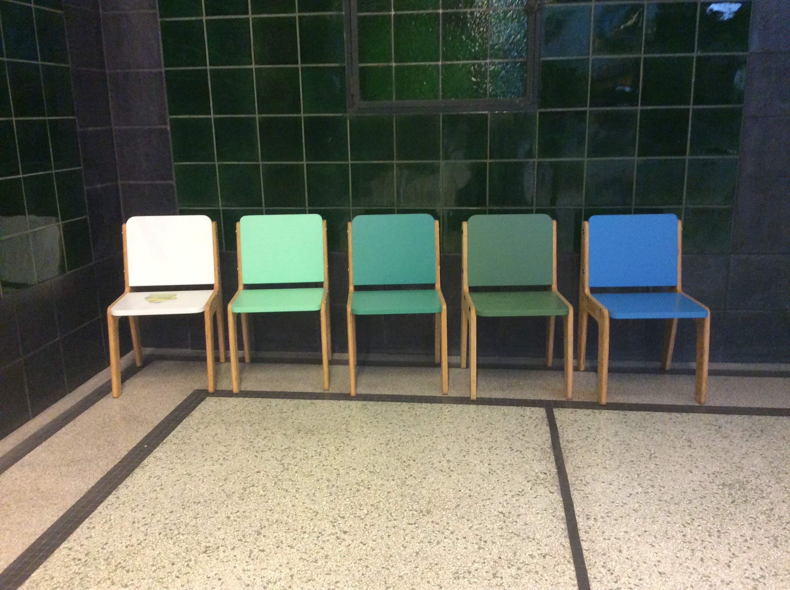 Vijf stoelen op een rij tegen een groene tegelwand, met zittingen in wit, mint, groen en blauw.